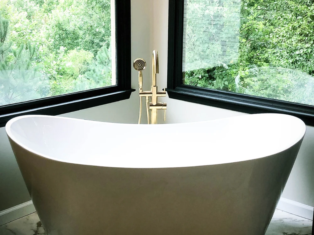 A bathroom with a freestanding soaking tub, a brass floor-mounted filler, and black-framed windows looking into trees.