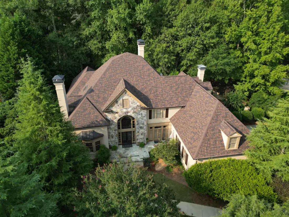 Aerial view of a large stone-and-brick house with a brown shingle roof, surrounded by mature trees.